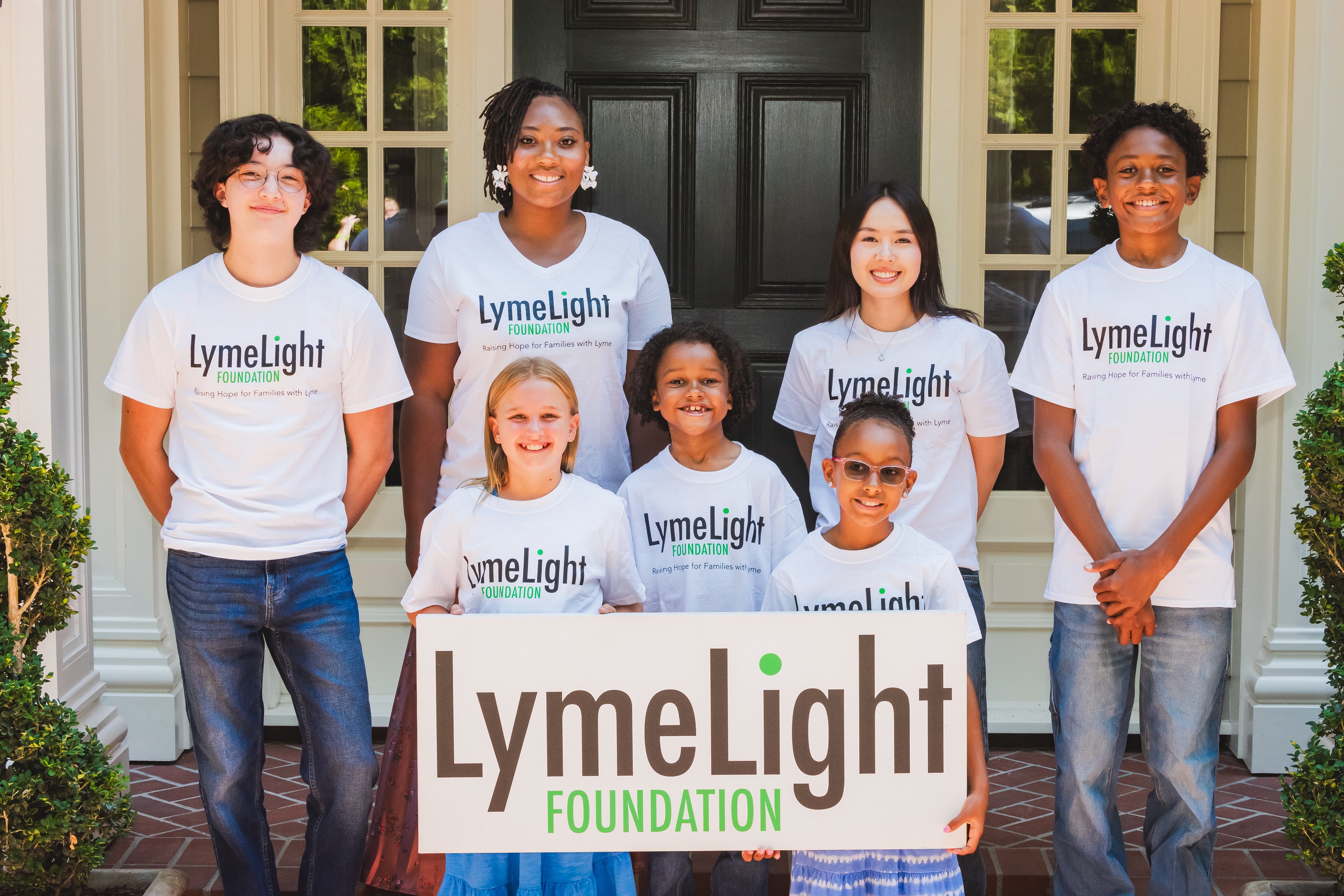Group of people wearing LymeLight t-shirts holding a LymeLight Foundation sign in front of a house.
