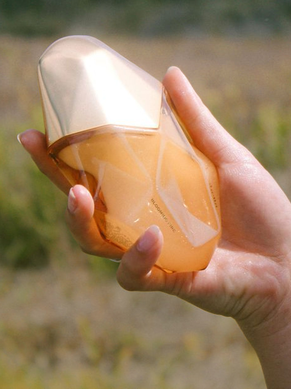 A hand holding a faceted, translucent amber-orange cosmetic bottle with a metallic gold cap, set against a blurred outdoor background.