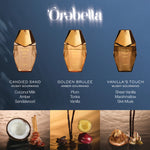 Three perfume bottles from Órabella are shown against a blue gradient background with scent names below each bottle: Candied Sand, Golden Brulee, and Vanilla's Touch. The bottom row features ingredient-style photos with coconut, spices, fruit, and vanilla elements.