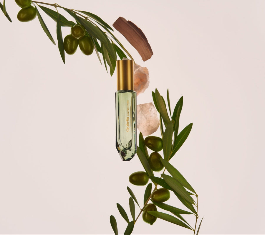 A clear glass roller bottle with a gold cap and gold lettering, suspended vertically between green olive branches and pale pink crystals against a solid light beige background.