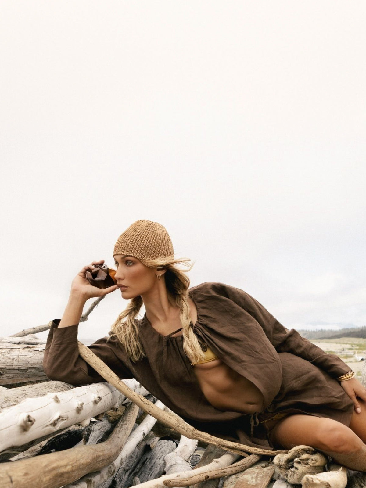 Bella Hadid lounging outdoors on driftwood on a beach, wearing a tan knit beanie, brown long-sleeve top, and gold jewelry while holding an Orebella Nightcap perfume bottle to her ear.