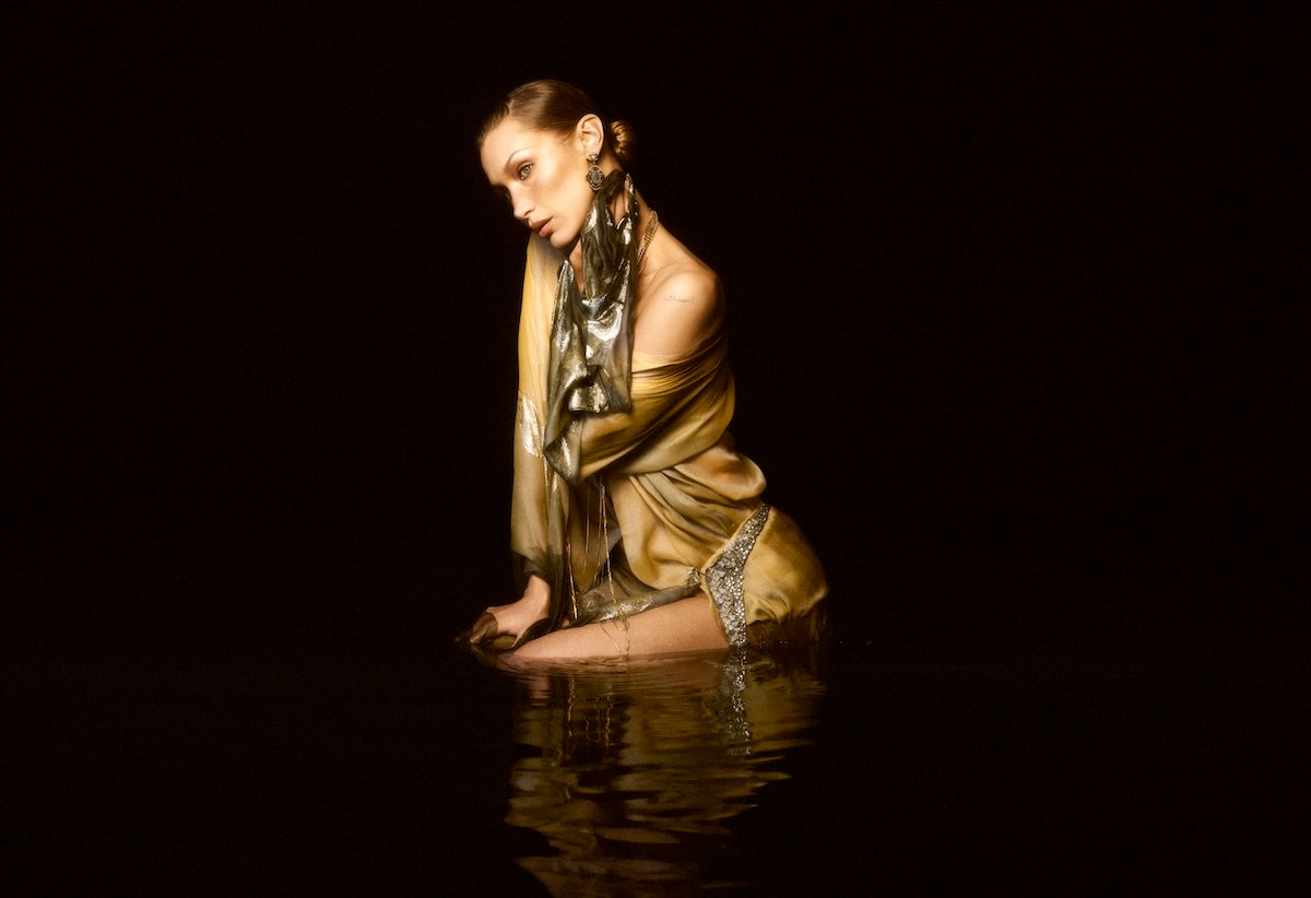 Bella Hadid kneeling in dark water against a black background, wearing a shimmering metallic gold dress with silver patterned trim and a matching metallic scarf.