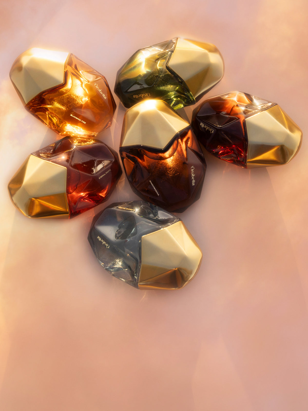 Six faceted perfume bottles arranged in a cluster on a soft peach-pink background. The bottles have metallic gold geometric tops and contain translucent amber, red, dark brown, and smoky gray liquids.