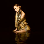 Bella Hadid kneeling in dark water against a black background, wearing a shimmering metallic gold dress with silver patterned trim and a matching scarf.