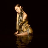 Bella Hadid kneeling in dark water against a black background, wearing a shimmering metallic gold dress with silver patterned trim and a matching scarf.