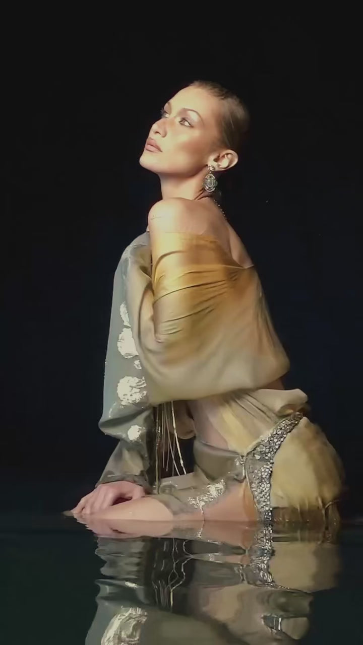 Bella Hadid posing gracefully in a sheer golden outfit while spraying Vanilla's Touch Body & Hair Perfume mist in campaign video 