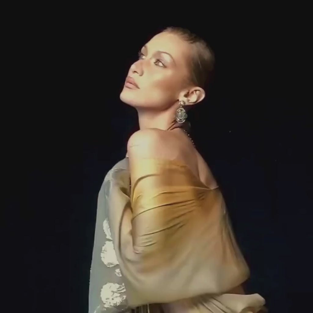 Bella Hadid posing gracefully in a sheer golden outfit while spraying Vanilla's Touch Body & Hair Perfume mist in campaign video 